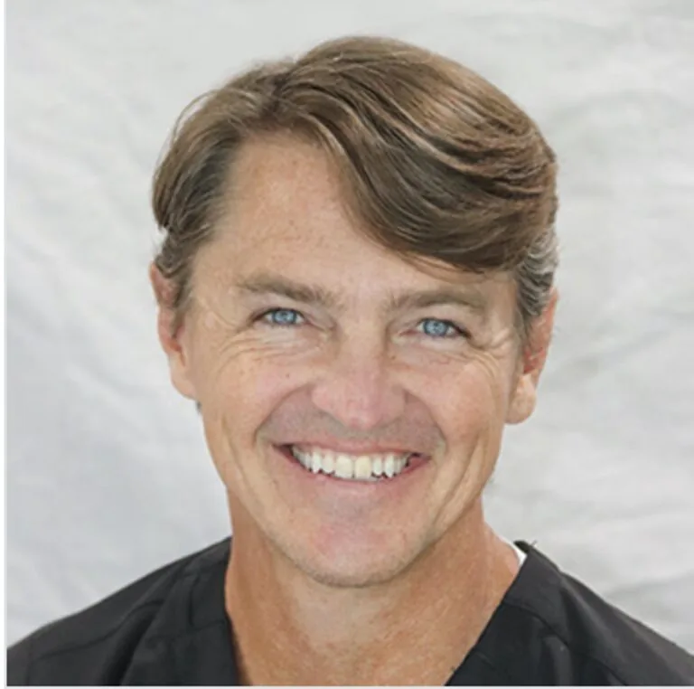 Person with short brown hair, blue eyes, black top, smiling broadly against a light background.