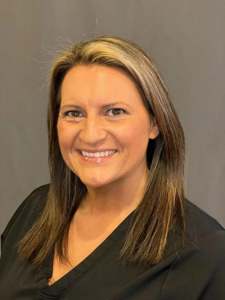 Portrait of a person with straight, shoulder-length brown hair featuring blonde highlights, wearing a black top and smiling directly at the camera. The background is a plain solid gray, keeping the focus on the subject’s face and expression.