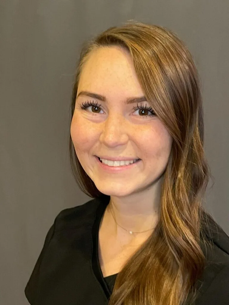 Portrait of a person with long, light brown hair styled in loose waves, wearing a black top and smiling directly at the camera. The background is a plain dark gray, creating a clean and professional visual focus.