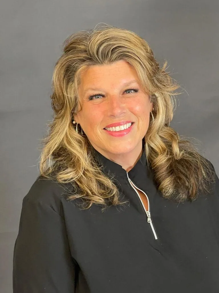 Portrait of a person with long, wavy hair featuring blonde highlights, wearing a black zip-up top and smiling directly at the camera. The background is a plain neutral gray, and the soft lighting emphasizes their facial features and hair texture.