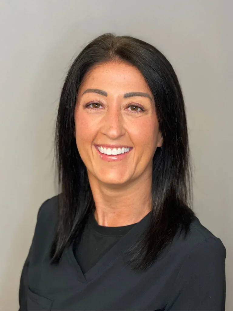 Portrait of a person with straight, shoulder-length black hair, wearing a black top and smiling warmly while looking directly at the camera. The background is a plain, light gray, emphasizing the subject’s face and expression.