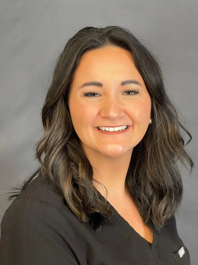 Portrait of a person with shoulder-length dark brown hair featuring lighter highlights, wearing a black top and smiling directly at the camera. The plain gray background keeps the focus on the individual’s face and expression.