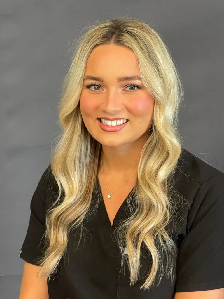 Professional portrait of a person with long, wavy blonde hair, wearing a black outfit and a small pendant necklace. They are seated against a plain dark gray background, smiling and looking directly at the camera.